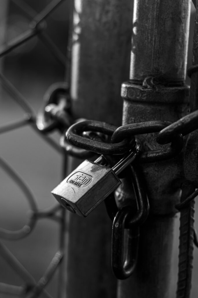 A detailed black and white image of a metal padlock securing a gate chain.